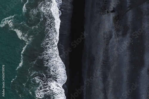 Fotografie Reynisfjara's black volcanic sand meets the turquoise Atlantic Ocean, with white waves creating striking contrasts and dynamic textures in Iceland
