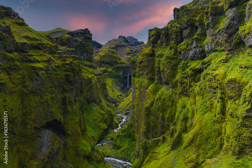 Fototapeta Naklejka Na Ścianę i Meble -  Mulagljufur Canyon in Iceland features moss covered cliffs, a winding river, and a distant waterfall under a pink and purple sunset sky.
