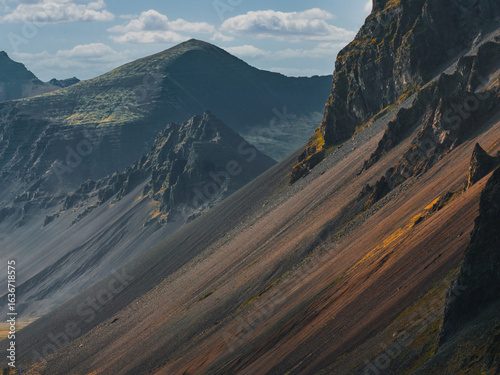 Steep volcanic slopes with dark rock and green patches under soft sunlight. Peaks rise in the background beneath a partly cloudy sky.