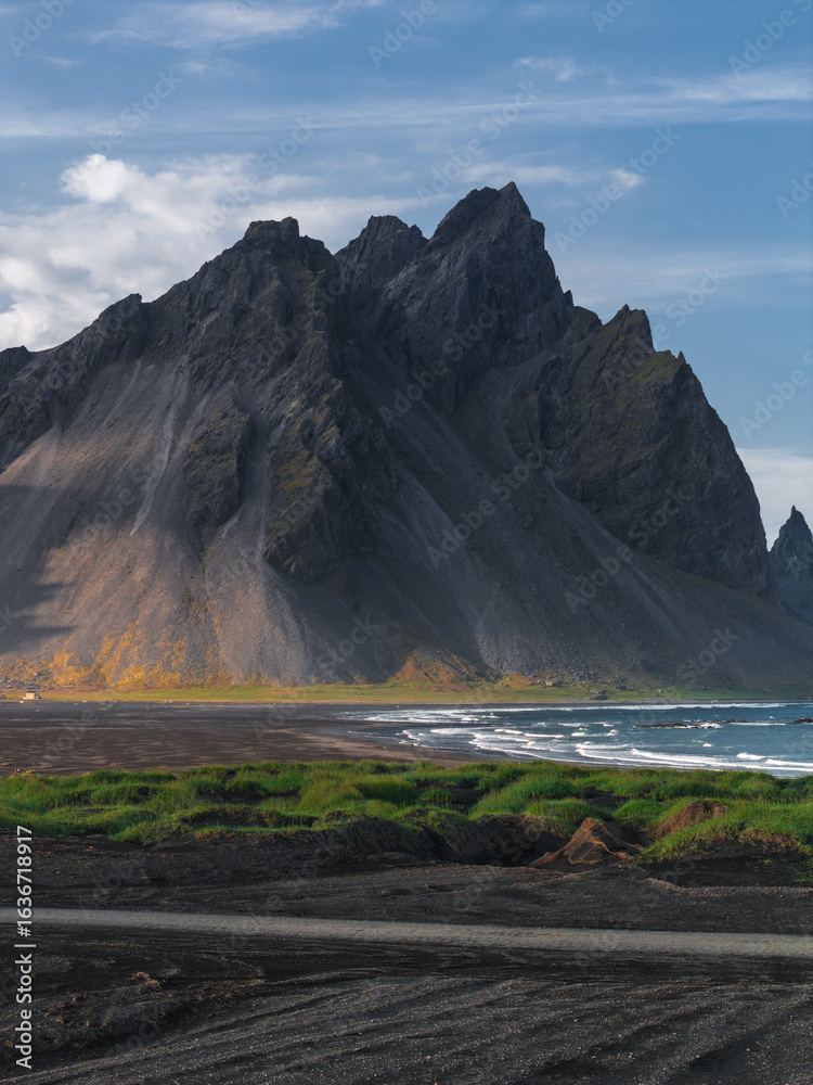 Fototapeta premium The Stokksnes Peninsula features Vestrahorn mountain's jagged peaks, a black sand beach with green grass, and ocean waves under a partly cloudy sky.