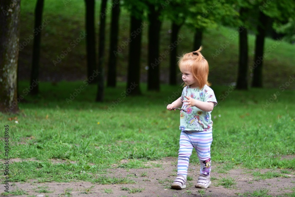 Red-haired toddler walks along grassy path near playground in sunny park, holding small stick. Red-haired toddler walks along grassy path near playground in sunny park, holding small stick.