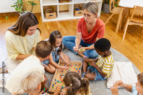 Bild auf Leinwand Preschool children and teachers sitting together, threading colorful beads to cr
