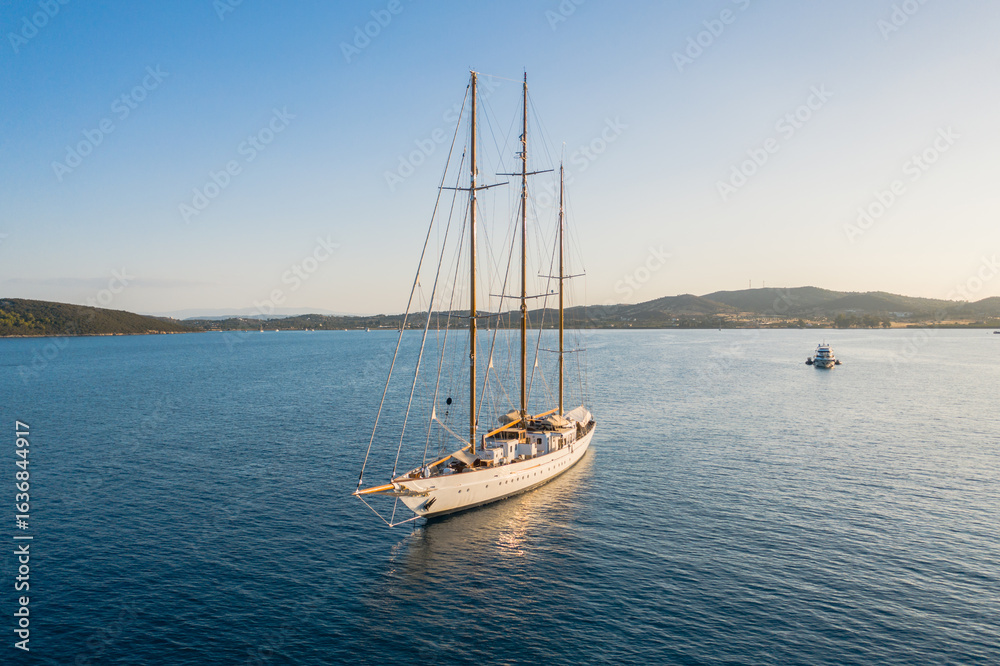 Aerial view of a majestic sailing yacht gliding across the tranquil, deep blue waters, kissed by the golden light of the setting sun, Ermioni, Argolis, Greece. Aerial view of a majestic sailing yacht gliding across the tranquil, deep blue waters, kissed by the golden light of the setting sun, Ermioni, Argolis, Greece.