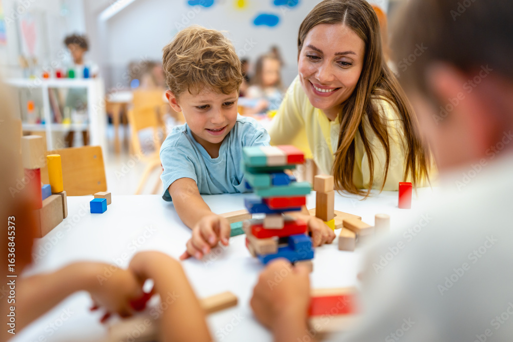 Fototapeta premium Smiling preschool boy playing with colorful wooden blocks, guided by a cheerful teacher, in a bright and lively classroom with other children in the background