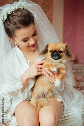 Bride in wedding veil sits on the bed holding a small fluffy dog, smiling at the camera. Tender and cozy atmosphere of the wedding morning.