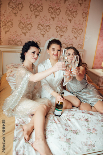 Bride in veil with bridesmaids sitting on the bed, raising glasses of champagne. Joyful, festive and friendly wedding morning atmosphere.