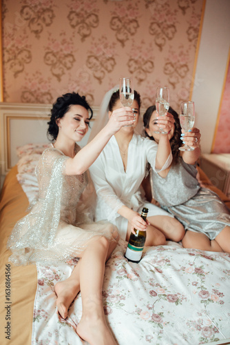 Bride in veil with bridesmaids sitting on the bed, raising glasses of champagne. Joyful, festive and friendly wedding morning atmosphere.