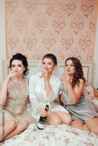 Bride in veil with bridesmaids sitting on the bed, raising glasses of champagne. Joyful, festive and friendly wedding morning atmosphere.