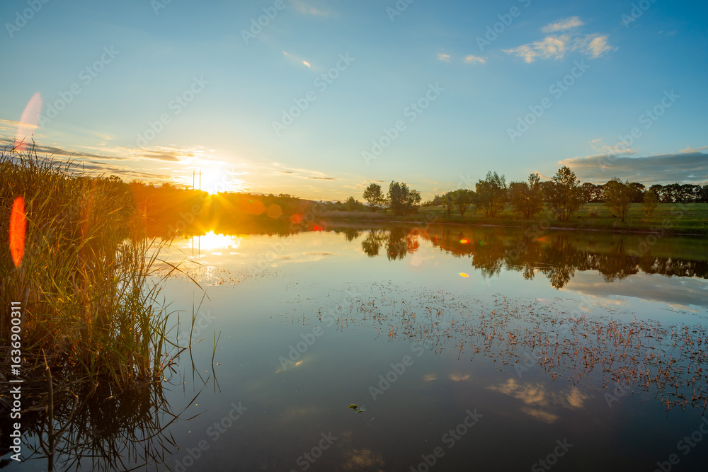 Fototapeta premium A tranquil evening view of a calm lake, with a bright sunburst and lens flares over the water, and a green shoreline with a distant treeline