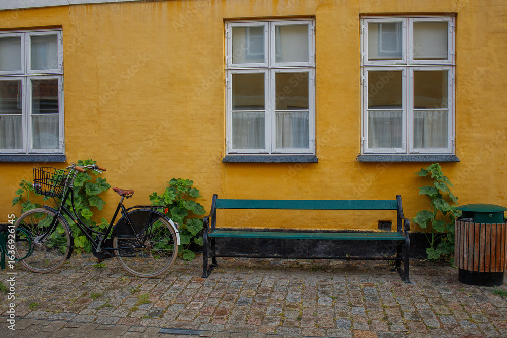 Small typical yellow old danish house in Dragor town. Bike parked near house, green bench and burdocks growing. Postcard from Denmark telling about danish lifestyle Small typical yellow old danish house in Dragor town. Bike parked near house, green bench and burdocks growing. Postcard from Denmark telling about danish lifestyle