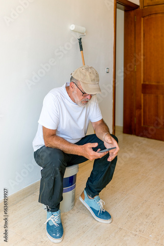 Senior painter sitting on paint bucket using smartphone during work break