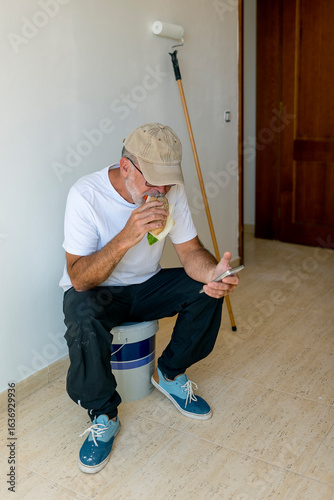 Painter sitting on paint bucket eating sandwich and using smartphone