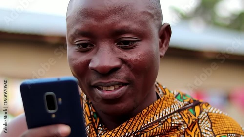 A happy African man wearing colorful traditional clothing smiles while using his smartphone outdoors
