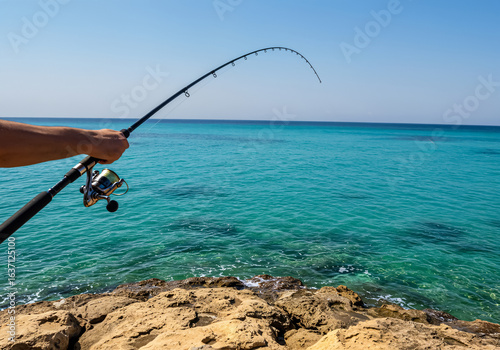 Person fishing from a rocky shore into clear turquoise mediterranean sea