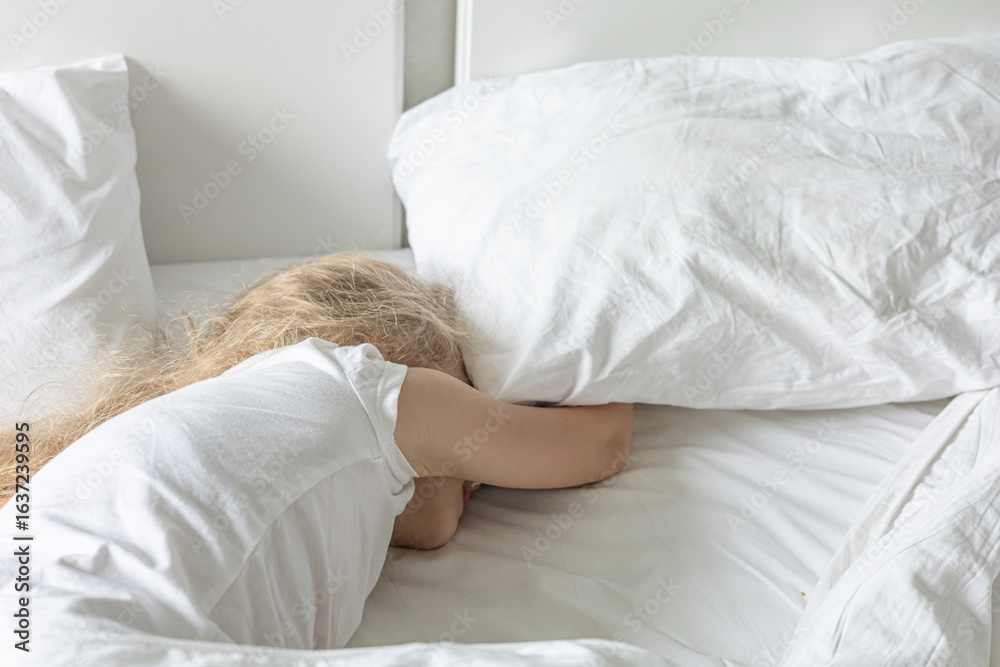 Blond child lying comfortably on a bed and hiding under the pillow. Child's Bedtime Routine, soft dreams. Waking up at weekends, lazy day Blond child lying comfortably on a bed and hiding under the pillow. Child's Bedtime Routine, soft dreams. Waking up at weekends, lazy day