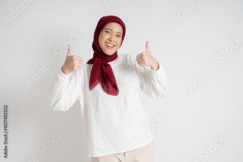 Confident woman expressing positivity with a thumbs up gesture while smiling warmly, featuring a vibrant red hijab and a light neutral background.