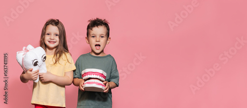 A smiling boy and a girl with healthy teeth hold a toy tooth in their hands on an isolated background. A place for your text. Oral hygiene. Pediatric dentistry. Prosthetics. Rules for brushing teeth.