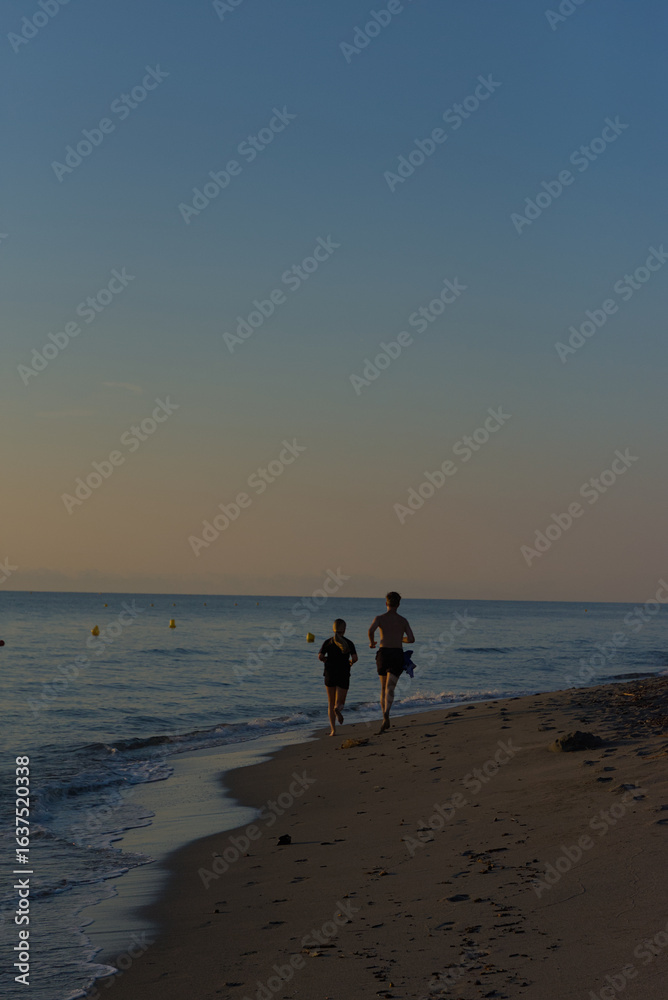 A young man and woman are seen jogging away from the camera on a sandy beach next to the ocean at sunset. A young man and woman are seen jogging away from the camera on a sandy beach next to the ocean at sunset.