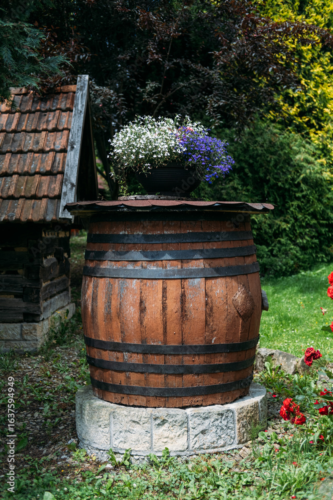 Naklejka premium Old wooden wine barrel repurposed as a garden planter with blooming flowers in a rustic outdoor setting. Upcycling, sustainable gardening, rural craftsmanship, traditional winemaking