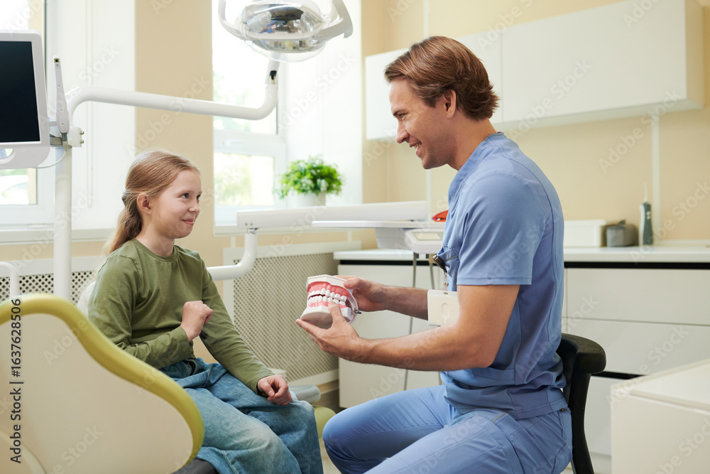 Obraz premium Caucasian male dentist showing dental model to Caucasian girl child sitting in dental chair, both smiling and interacting in modern dental clinic with equipment visible