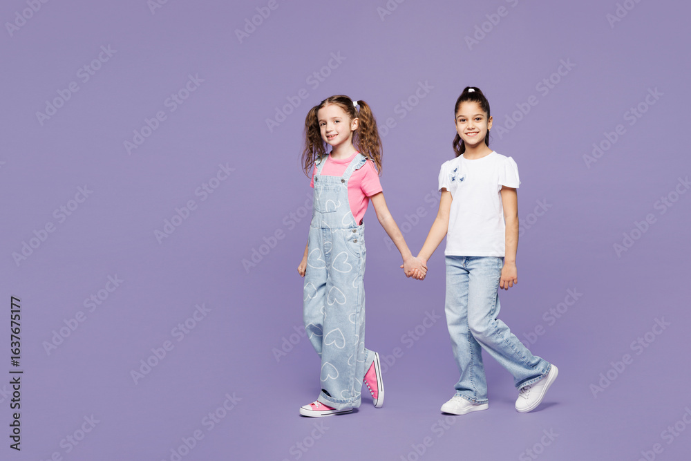 Obraz premium Full body side profile view little two friends kids children girls 9-10 years old wearing casual clothes together hold hands walk go look camera isolated on plain purple background. Lifestyle concept.