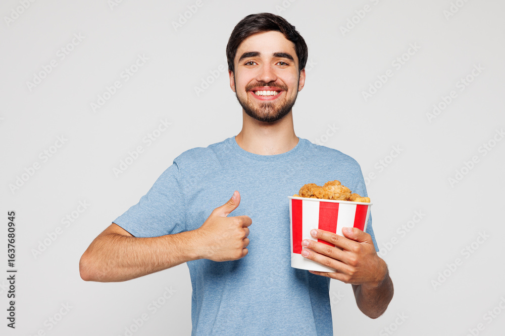 Young man wears blue casual clothes hold takeaway box with barbecue chicken meat wings show thumb up isolated on plain solid white background. Proper nutrition healthy food unhealthy choice concept. Young man wears blue casual clothes hold takeaway box with barbecue chicken meat wings show thumb up isolated on plain solid white background. Proper nutrition healthy food unhealthy choice concept.