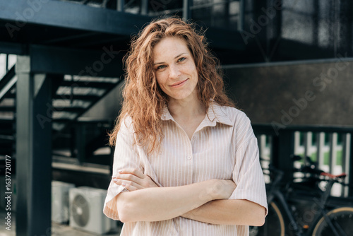 A beautiful confident woman stands with her arms crossed against the background of a modern city building.