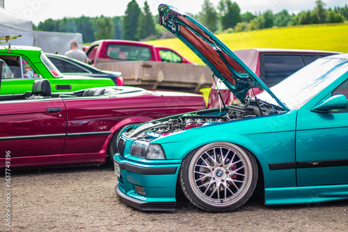 Custom cars with vibrant paint, lowered suspensions, and open hoods displayed at an outdoor automotive tuning show on a sunny day.