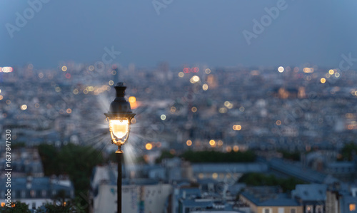 Fototapeta Naklejka Na Ścianę i Meble -  Parisian street lamp glows warmly against a blurred backdrop of the city lights at dusk, creating a romantic and nostalgic atmosphere