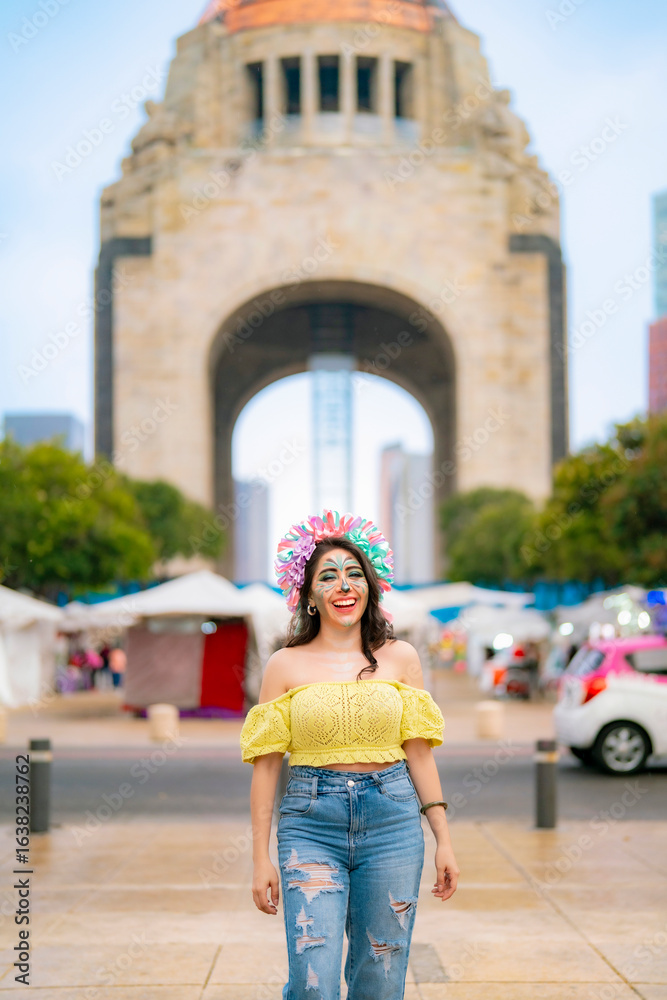 Fototapeta premium Smiling young Latina with colorful catrina makeup walks at Monumento a la Revolucion in Mexico City during Dia de Muertos, wearing yellow top and ripped blue jeans, radiating confidence and tradition