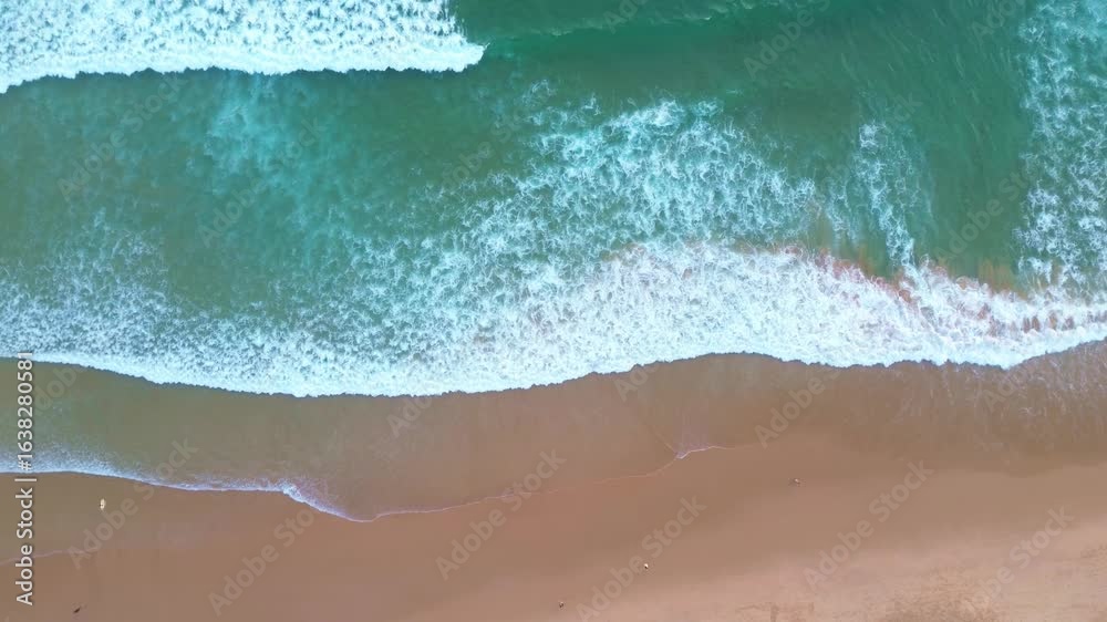 Overhead drone view of a sandy beach with gentle blue-turquoise waves rolling onto the shore, surfers walking along the coastline, ocean at the end of the day before evening.