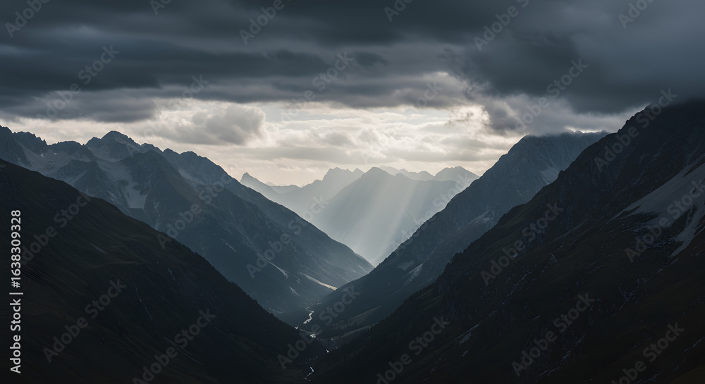 Obraz premium Epic vista of a mountain valley shrouded in moody clouds and pierced by radiant light streams, evoking a sense of vastness and serene grandeur with peaks layered in atmospheric perspective