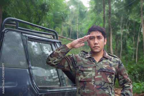 An Asian army officer in uniform salutes directly at the camera, deep in a lush jungle with an off-road vehicle visible in the background