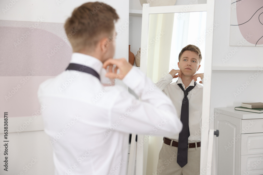 Fototapeta premium Getting ready. Young man tying stylish necktie near mirror at home