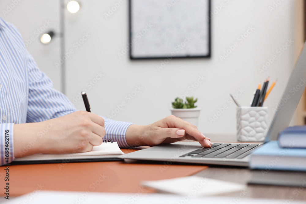 Fototapeta premium Copywriter using laptop and writing in notebook at wooden table indoors, closeup