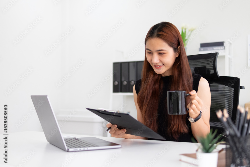 A businesswoman is sitting in an office doing paperwork, she is staring at a clipboard and using a laptop on the table. A businesswoman is sitting in an office doing paperwork, she is staring at a clipboard and using a laptop on the table.