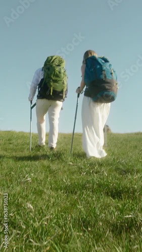 In a vertical video, two adventurous wedding couples dressed in wedding outfits walk together across open grassy hillsides with heavy mountain backpacks beneath bright blue skies