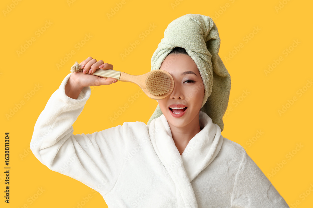 Young Asian woman in bathrobe with massage brush after shower on yellow background Young Asian woman in bathrobe with massage brush after shower on yellow background