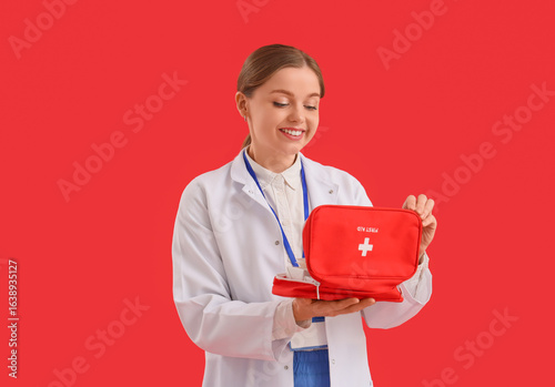 Young female doctor with first aid kit on red background