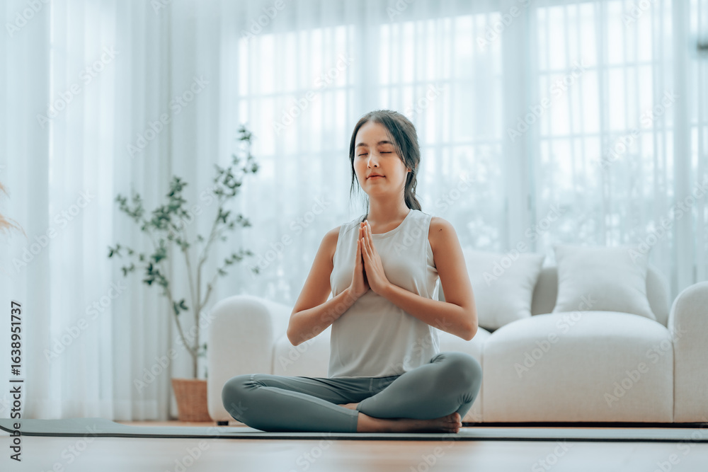 Young Asian woman meditating in lotus position at home, sitting on the floor in fitness clothing. Breathing workout to achieve relaxation, peace and mindfulness Young Asian woman meditating in lotus position at home, sitting on the floor in fitness clothing. Breathing workout to achieve relaxation, peace and mindfulness