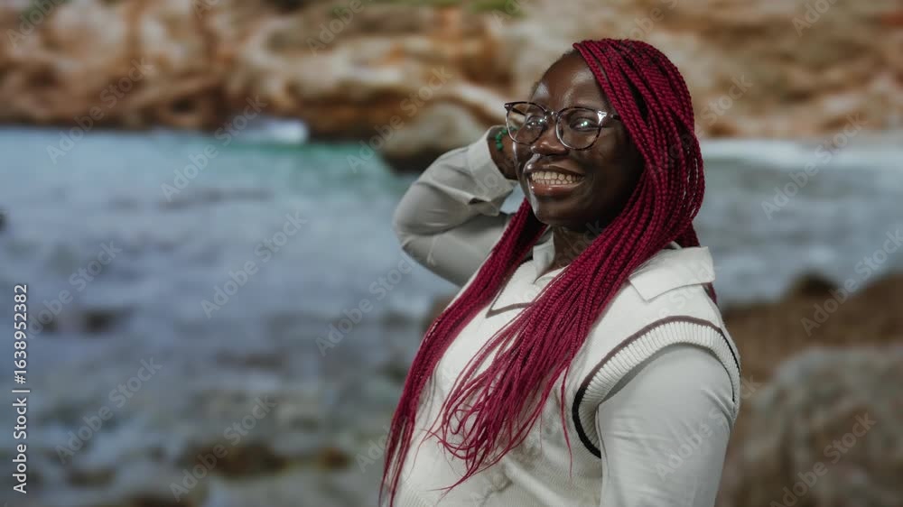 Woman smiling in front of a scenic seaside with vibrant pink braids and glasses, capturing a joyful outdoor beach moment with a rocky backdrop and casual attire.