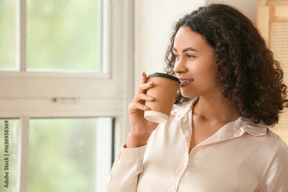 Beautiful young African-American businesswoman with paper cup of hot coffee near window in office Beautiful young African-American businesswoman with paper cup of hot coffee near window in office