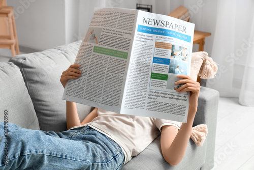 Young woman reading newspaper on grey sofa at home