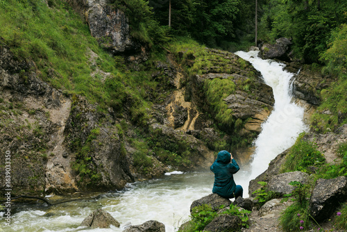 A person in a teal jacket photographing the powerful Kuhflucht Gorge waterfall in Farchant, Bavaria, Germany. The cascading water flows through moss-covered rocks and lush green forest, capturing the 