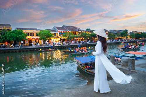 Asian woman wearing traditional Vietnamese cultural dress in Hoi An Ancient Town, Hoi An, Vietnam at dusk with colorful lights from boats.