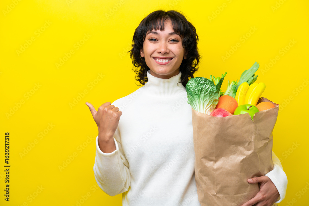 Young Argentinian woman holding a grocery shopping bag isolated on yellow background pointing to the side to present a product Young Argentinian woman holding a grocery shopping bag isolated on yellow background pointing to the side to present a product