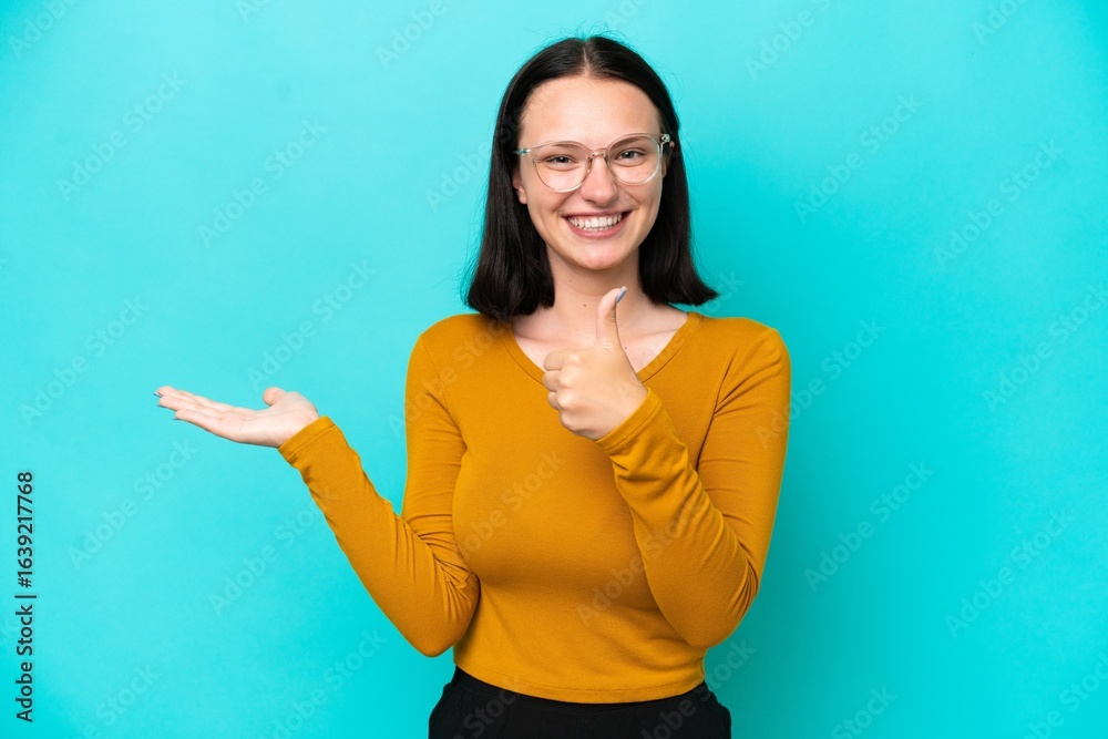 Fototapeta premium Young caucasian woman isolated on blue background holding copyspace imaginary on the palm to insert an ad and with thumbs up