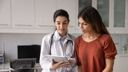 Optimistic young Hispanic doctor woman showing medical report on tablet to female patient sitting on couch, telling checkup result, giving prescription, treatment advice. Banner shot