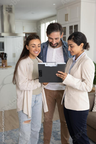 Real estate agent showing document to couple, discussing details of property, reviewing listing or contract, standing in modern flat. Designer demonstrate interior design ideas to clients on clipboard