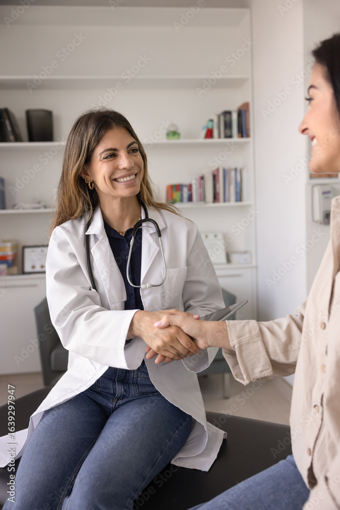 Fototapeta premium Happy young Latin doctor woman giving handshake to positive female patient, wishing getting better after examination, medical checkup, consultation, sitting on couch, smiling